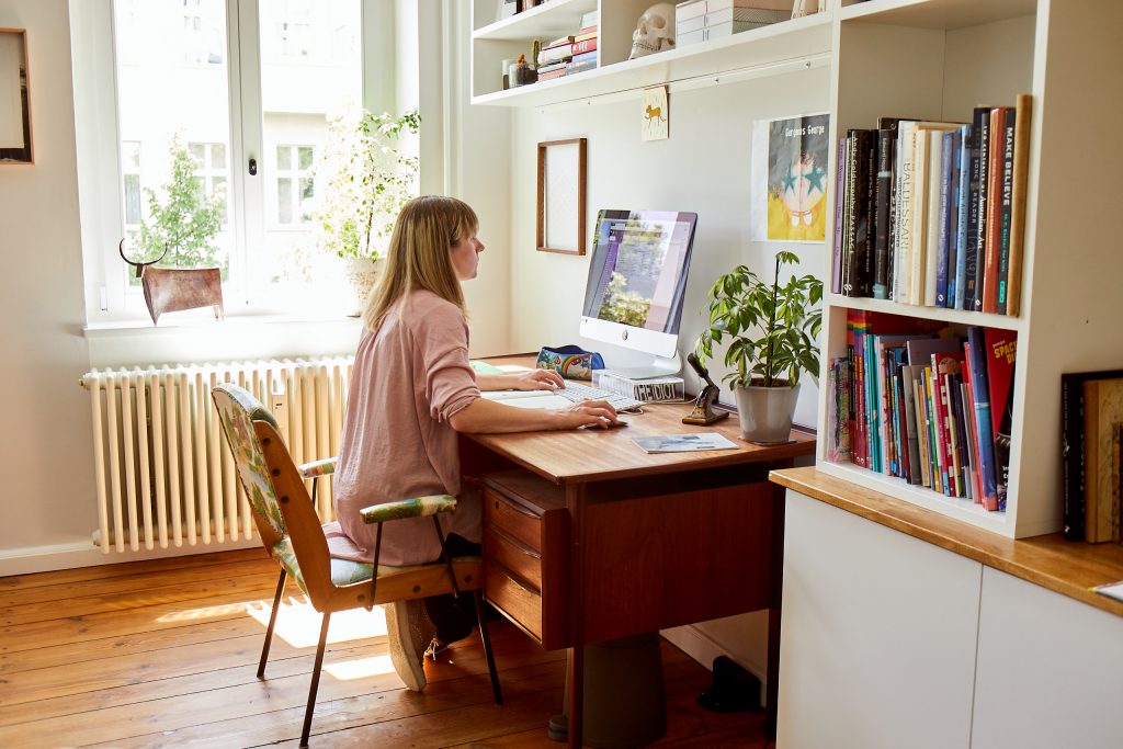 Kristen Harrison sits at her desk in the office of the Curved House. She is at a computer surrounded by books and plants. 